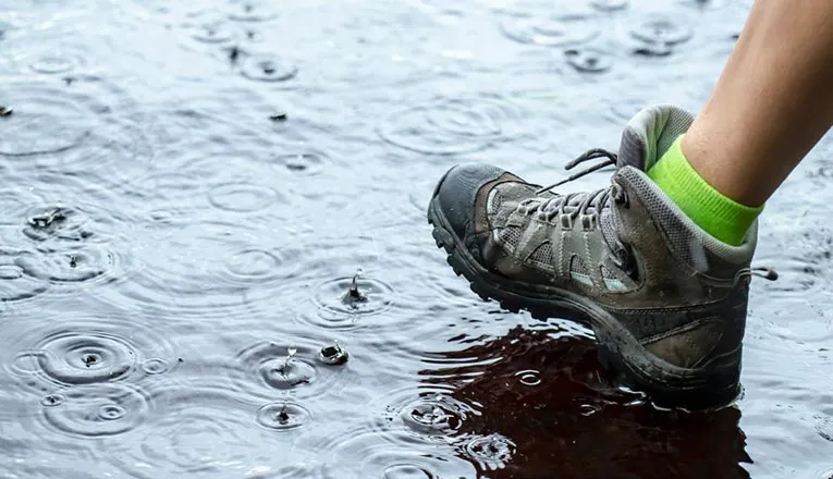 A person's foot, clad in a hiking boot, is stepping into a puddle, creating ripples on the water's surface.